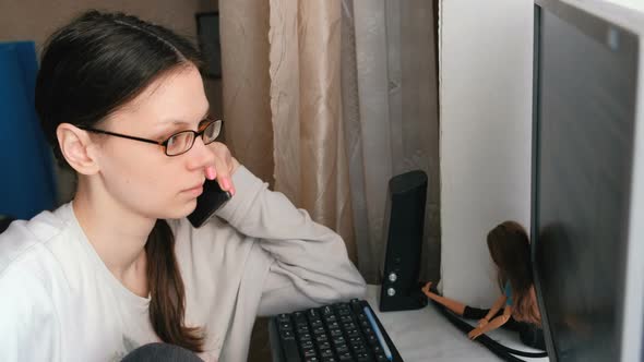 Young Woman in Glasses Working at Computer and Speaking Mobile Phone.