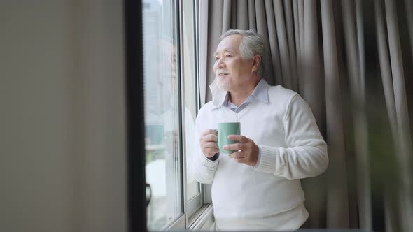 asian male white beard stay home hand hold hot drink standing look outside of apartmen