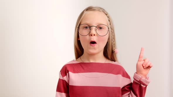 Little clever girl child in glasses looks at the camera amazed raising a finger up an idea alt