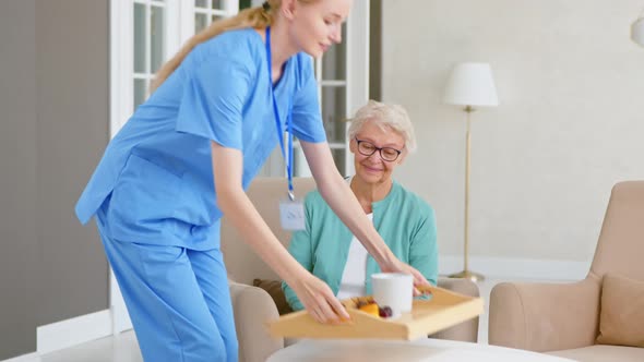Lady medical worker in blue uniform carries drink and fruits to old woman alt