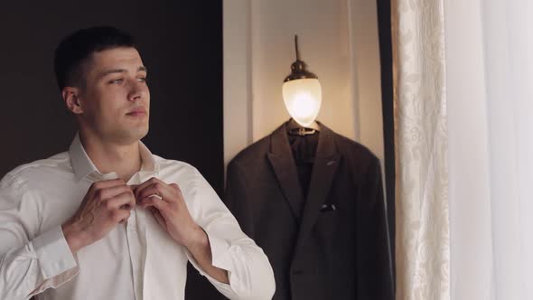 Stylish Man in Jacket Fastens Buttons Buttoning White Shirt Preparing to Go Out Near Window alt