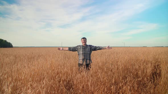 A farmer stands in a field with wheat spreads his arms to the sides.