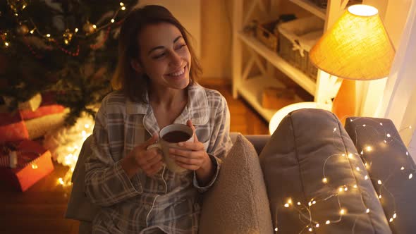 Happy Dreaming Girl in Pajama Sitting on Couch and Holding Hot Chocolate Mug Drinking and Relaxing alt