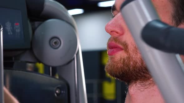 A Young Fit Man Trains on a Machine in a Gym - Face Closeup From the Side alt