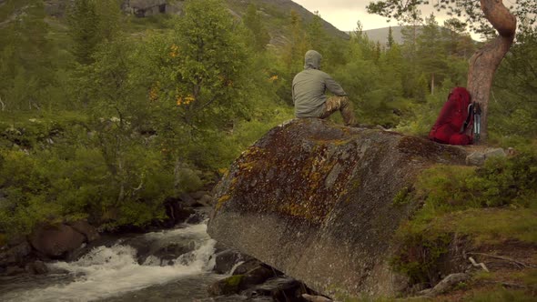 A Traveler with a Red Backpack Sitting on the Rock Above the Mountain Waterfall alt