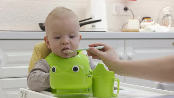 Teaching Infant Baby to Self Feed Toddler in Green Bib on Feeding High Chair Trying to Eat Porridge alt