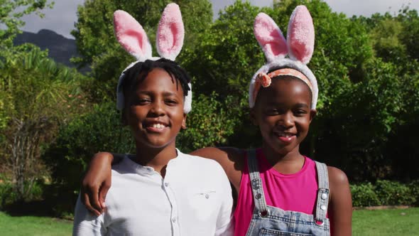 Smiling african american brother and sister wearing easter bunny ears embracing in garden alt