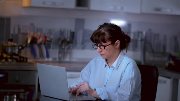 Pretty Woman with Glasses Sits at a Laptop in the Kitchen at Home