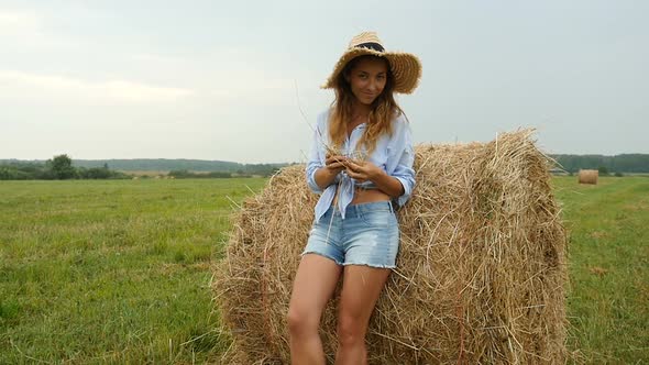 Pretty Young Woman Stands Near Haystack and Plays With Straw. Farmer's Wife Walks on the Field