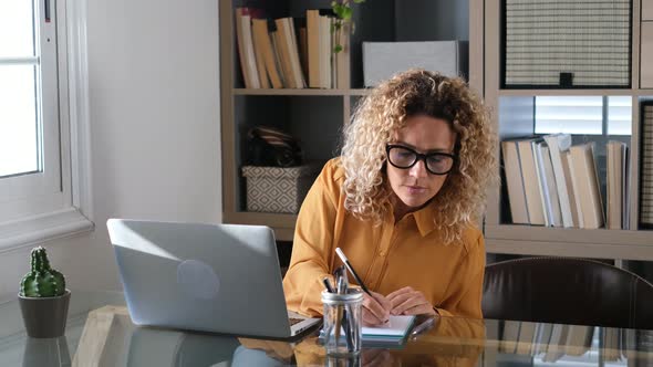Blond businesswoman writing notes sitting at desk with laptop