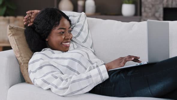 Smiling African Woman Student Girl Freelancer User Lying on Couch Using Laptop Typing Message Online alt