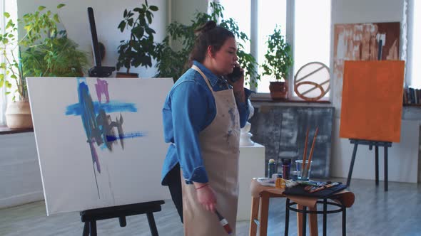 A Woman Paint Artist Talking on Her Phone in the Studio in the Middle of the Drawing Process alt
