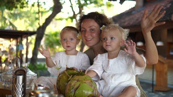 Portrait of Young Mom with Her Twin Daughters in the Beach Cafe Drinking Coconut Milk Waving to the alt