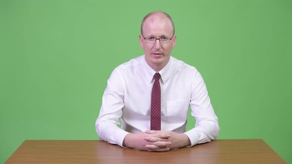 Mature Bald Businessman Looking Tired While Explaining Something Against Wooden Table alt