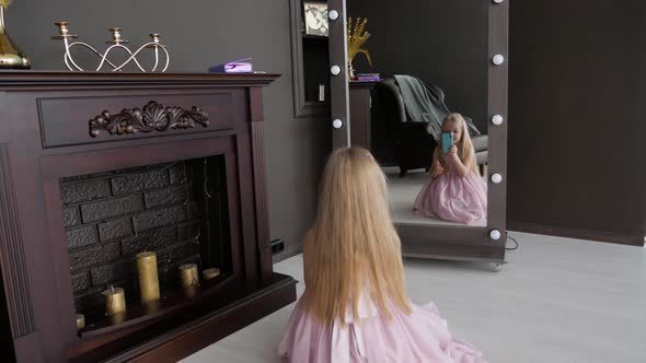 Little girl with blond hair sitting on floor in dark room in front of mirror taking selfie photo