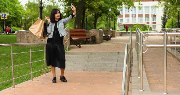 Brunette woman holding paper shopping bags and taking selfie by smartphone in park alt
