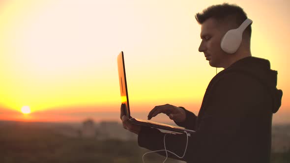 A Man Freelancer in Headphones Standing on the Roof at Sunset Writes on the Keyboard Code Pages alt