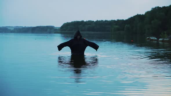Evil witch in black cloak with hood in the river. Halloween costume. alt