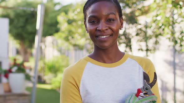 Portrait of african amercian woman holding secateur looking to camera and smiling in garden alt