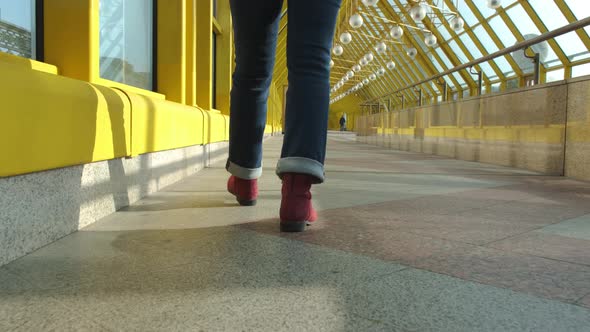 Women's Legs in Burgundy Boots and Blue Jeans Walk on a Closed Pedestrian Bridge alt
