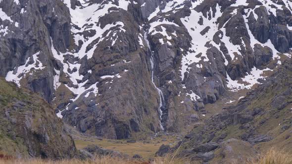 Static, hiker walks past distant waterfall in snow capped landscape, Routeburn Track New Zealand alt