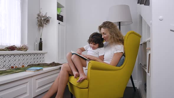 Mom Reads Child book to her little son while sitting in comfortable yellow armchair alt
