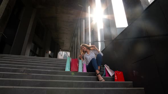 Teenager Smiling Girl with Shopping Bags Sitting on Stairs Near Shopping Mall. Black Friday Sale alt