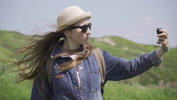 A Woman with Long Hair in a Hat and Sunglasses is Filming Herself in Nature alt