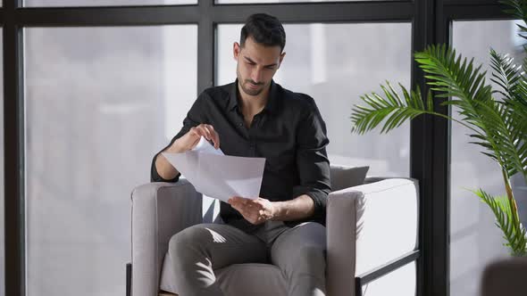 Portrait of Confident Young Man Sitting at Window in Armchair Analyzing Documents alt
