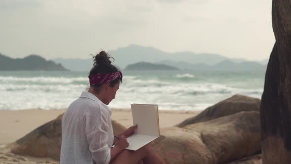 Brunette Woman Sits on Wild Beach Writing in Notebook