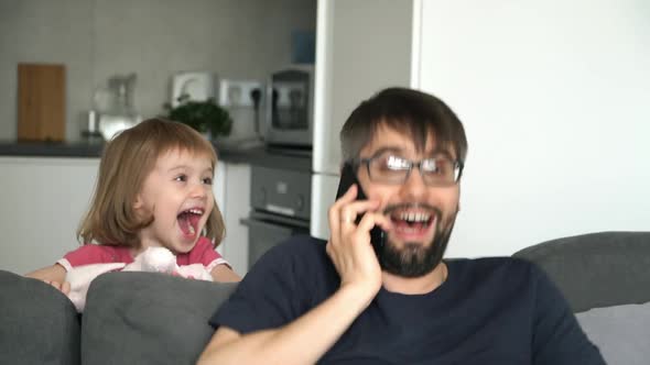 Child Girl Jumps Out From Behind Sofa, Shout, Laughs and Scares Father Talking on Phone. alt