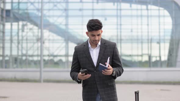 Likable Young Indian Man Using Mobile Phone Standing Outside Airport Terminal Holding Passport alt