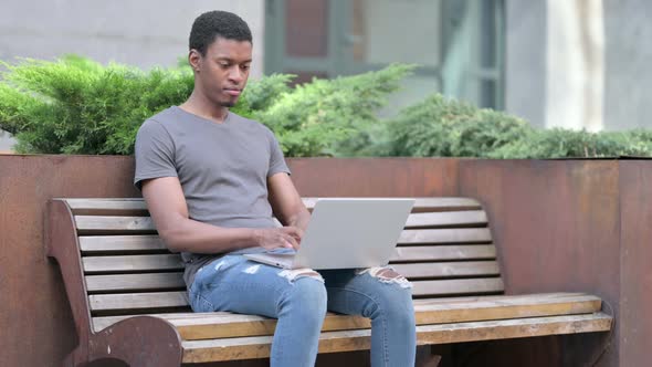 Focused Young Young African Man Using Laptop on Bench  alt