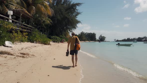 A man walks barefoot along the beach with his shoes in his hands and a satchel over his shoulder alt