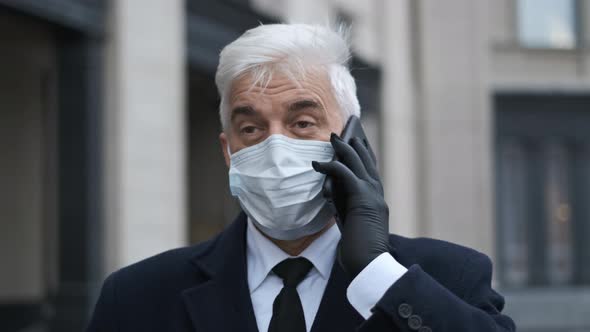 Senior Masked Businessman with Gray Hair in Black Coat Gloves Speaks on Phone alt