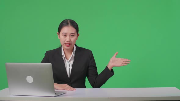 Live News Studio With Asian Female Anchor And Her Computer Pointing While Reporting On Green Screen alt