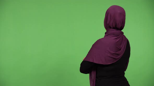 A Muslim Woman Looks at the Green Screen Background with Her Back to the Camera