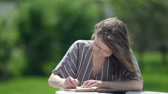 a Brunette in a Striped Dress and Glasses is Sitting at a Table Outside on a Sunny Day Against a alt
