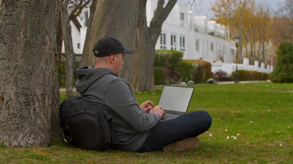 Young Man is Sitting in a Park in a Tourist Place Working on a Laptop alt