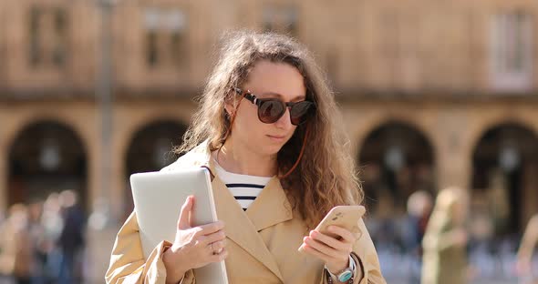 Smiling young woman wearing relax outfit using mobile phone typing text messages walking holding lap