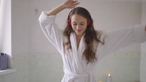 Medium Shot of Joyful Young Caucasian Slim Woman in Bathrobe and Headphones Dancing in Slow Motion alt