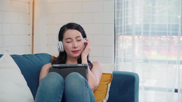 Asian woman listening music and using tablet, female using relax time lying on home sofa. alt