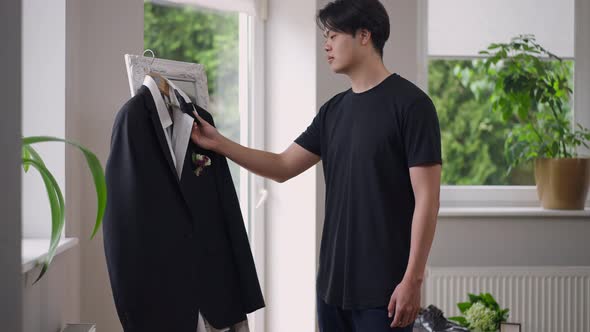 Smiling Happy Asian Groom Admiring Elegant Blazer with Bow Tie Hanging on Mirror Indoors alt