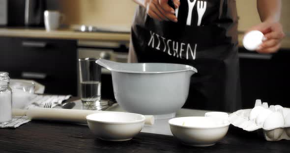 Woman in an Apron Breaks an Egg Into a Bowl with a Knife in a Modern Kitchen alt