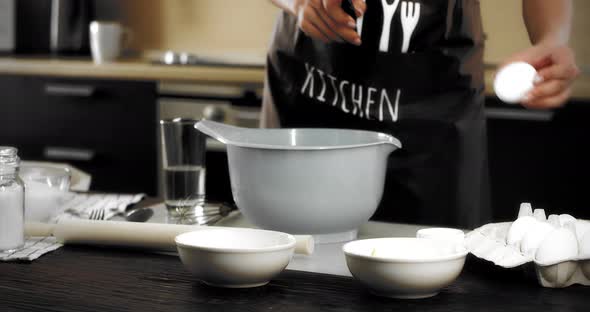 Woman in an Apron Breaks an Egg Into a Bowl with a Knife in a Modern Kitchen