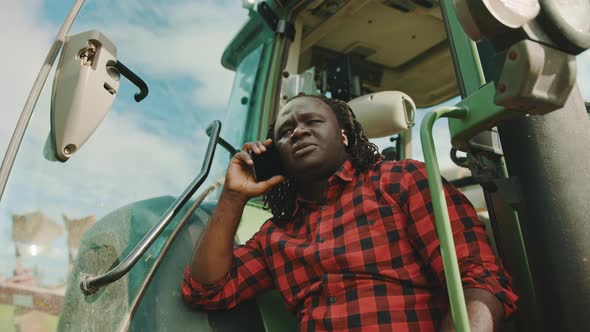 Young African Farmer Speaking on the Smartphone While Leaning on the Green Tractor alt