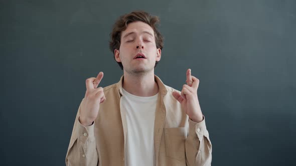 Portrait of Hopeful Man Crossing Fingers and Closing Eyes Praying Standing on Gray Color Background alt