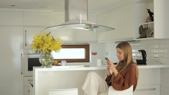 Young blond woman using smartphone and drinks tea in the white kitchen in good mood alt