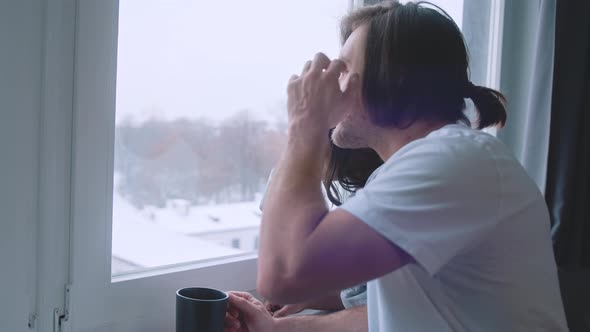 Happy Multiracial Couple Drinking Hot Beverage While Looking Through the Window at First Snow alt