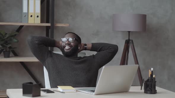 Relaxed African Man Wireless Headphones Enjoy Listen Music Hand Behind Head Sit at Work Desk Young alt